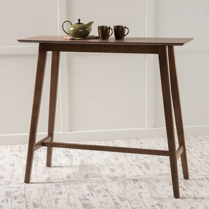 A Bar Height Dining Table With Splayed Legs And Elongated Rectangular Top stands on a white textured floor. On the tabletop are a green teapot with a lid and two matching ceramic cups. White paneled walls form the background.