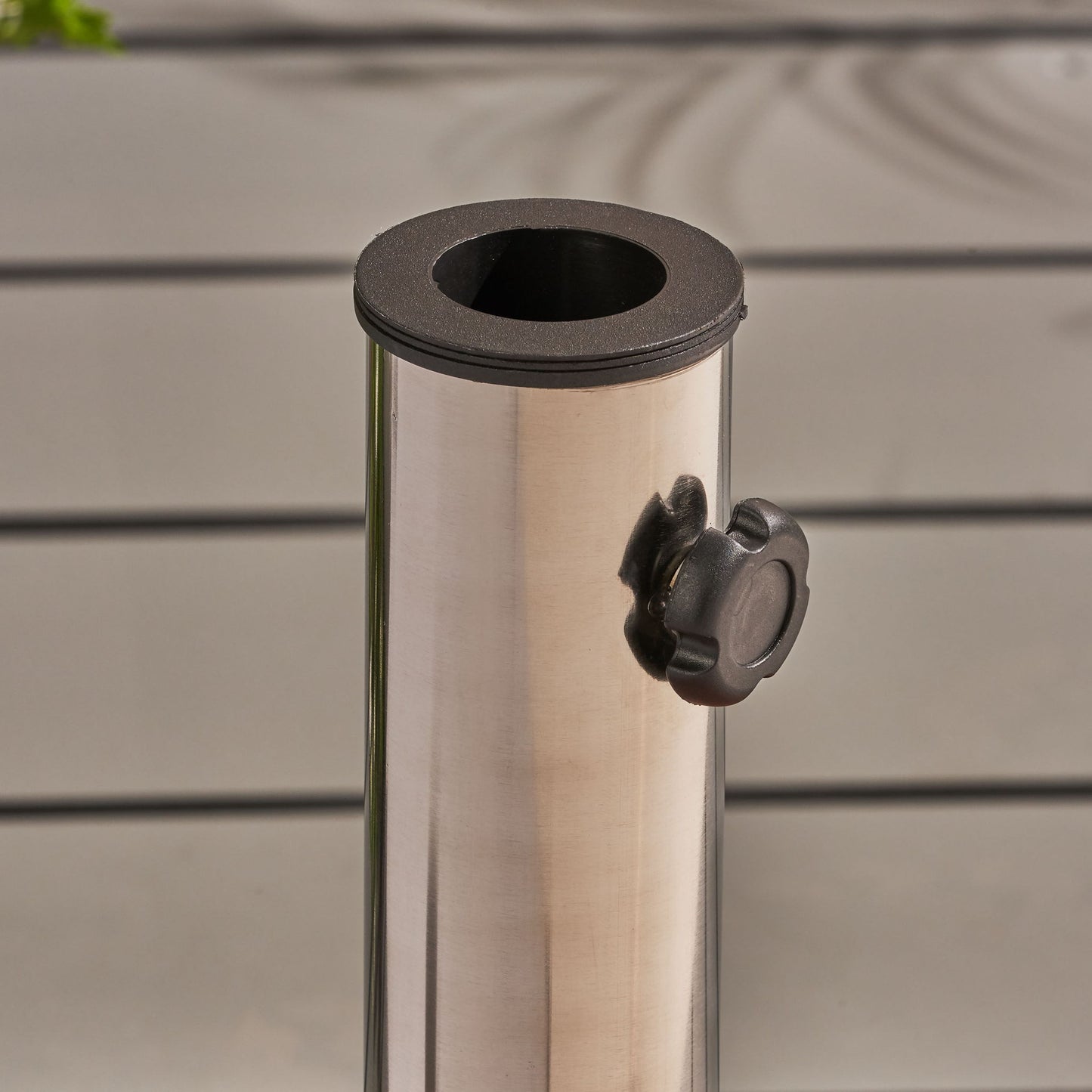 A close-up of the Wheelie - Round Umbrella Base With Stainless Steel Holder featuring a black plastic tightening knob, set against a light gray slatted background.