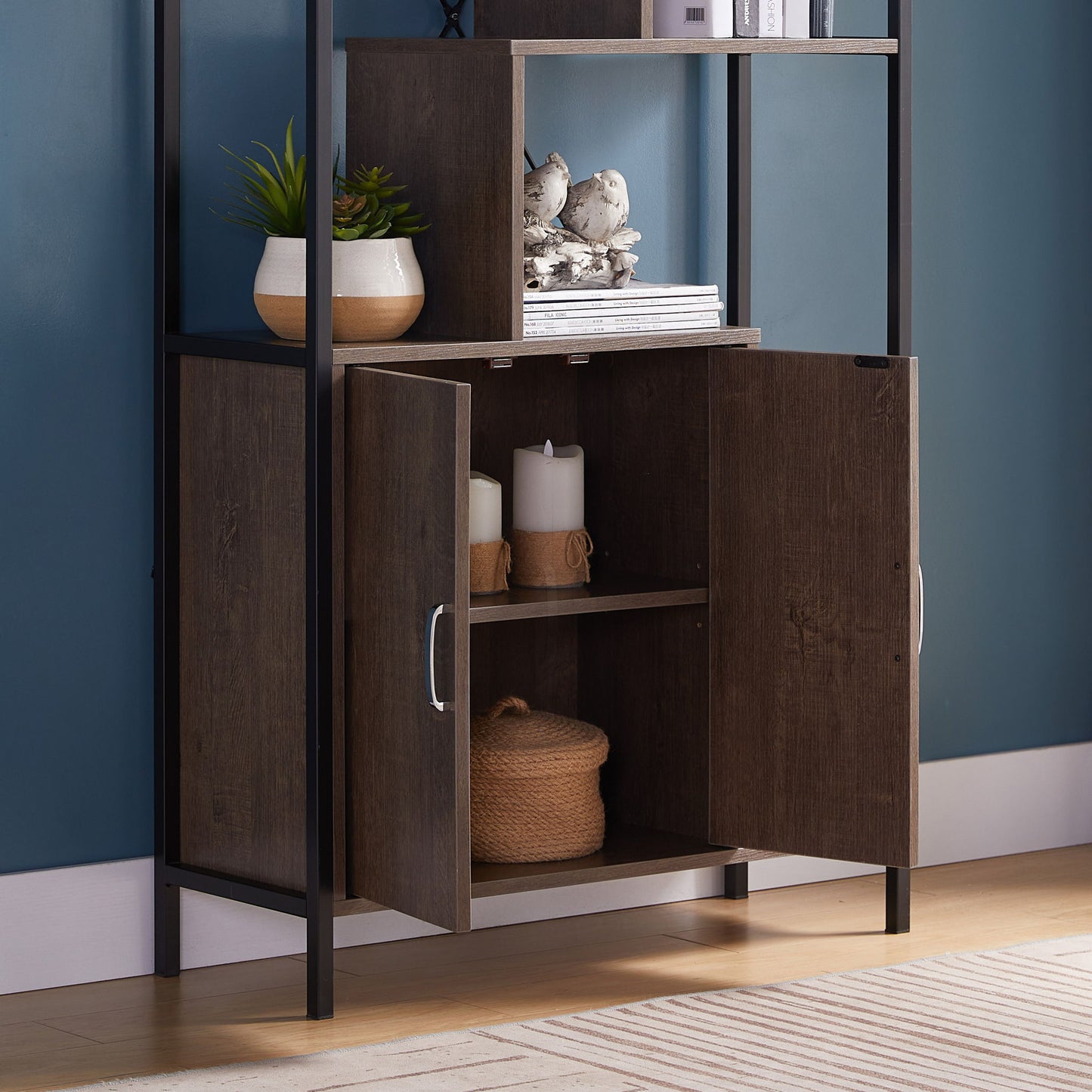 The Modern Bookcase With Two Door Storage Cabinet With Two Shelves, featuring a black metal frame, stands against a blue wall with its doors open to reveal candles and a woven basket. Potted plants and bird figurines decorate the top shelf.