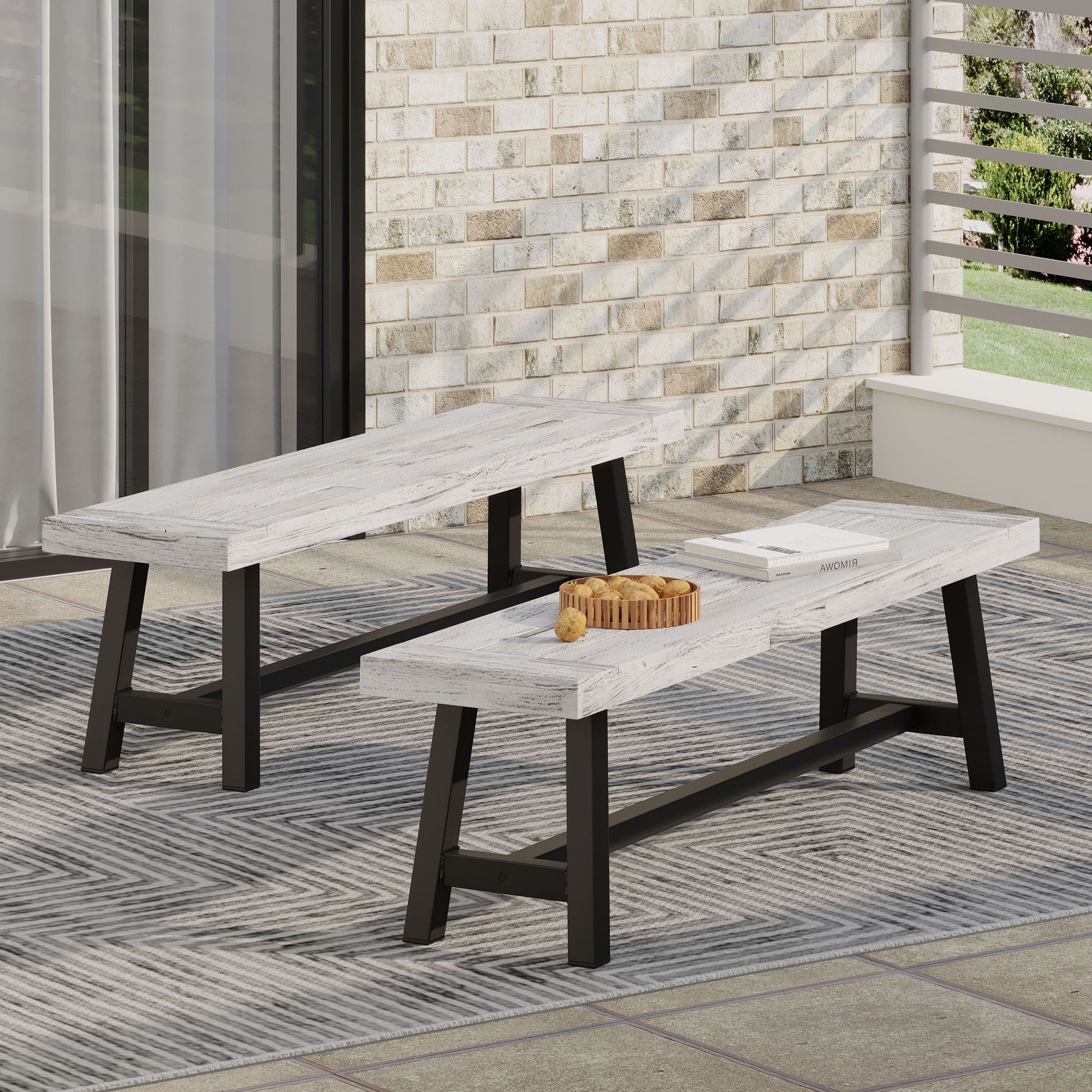 Two Tranquilizer Outdoor Acacia Wood Benches with slat panel design and black metal legs sit on a patterned rug on the patio. One bench holds a wooden tray with drinks and fruit, against a brick wall and railing backdrop.
