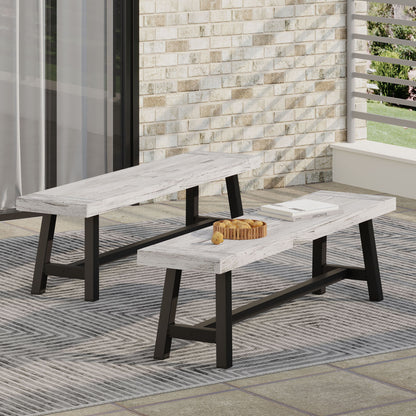 Two Tranquilizer Outdoor Acacia Wood Benches with slat panel design and black metal legs sit on a patterned rug on the patio. One bench holds a wooden tray with drinks and fruit, against a brick wall and railing backdrop.