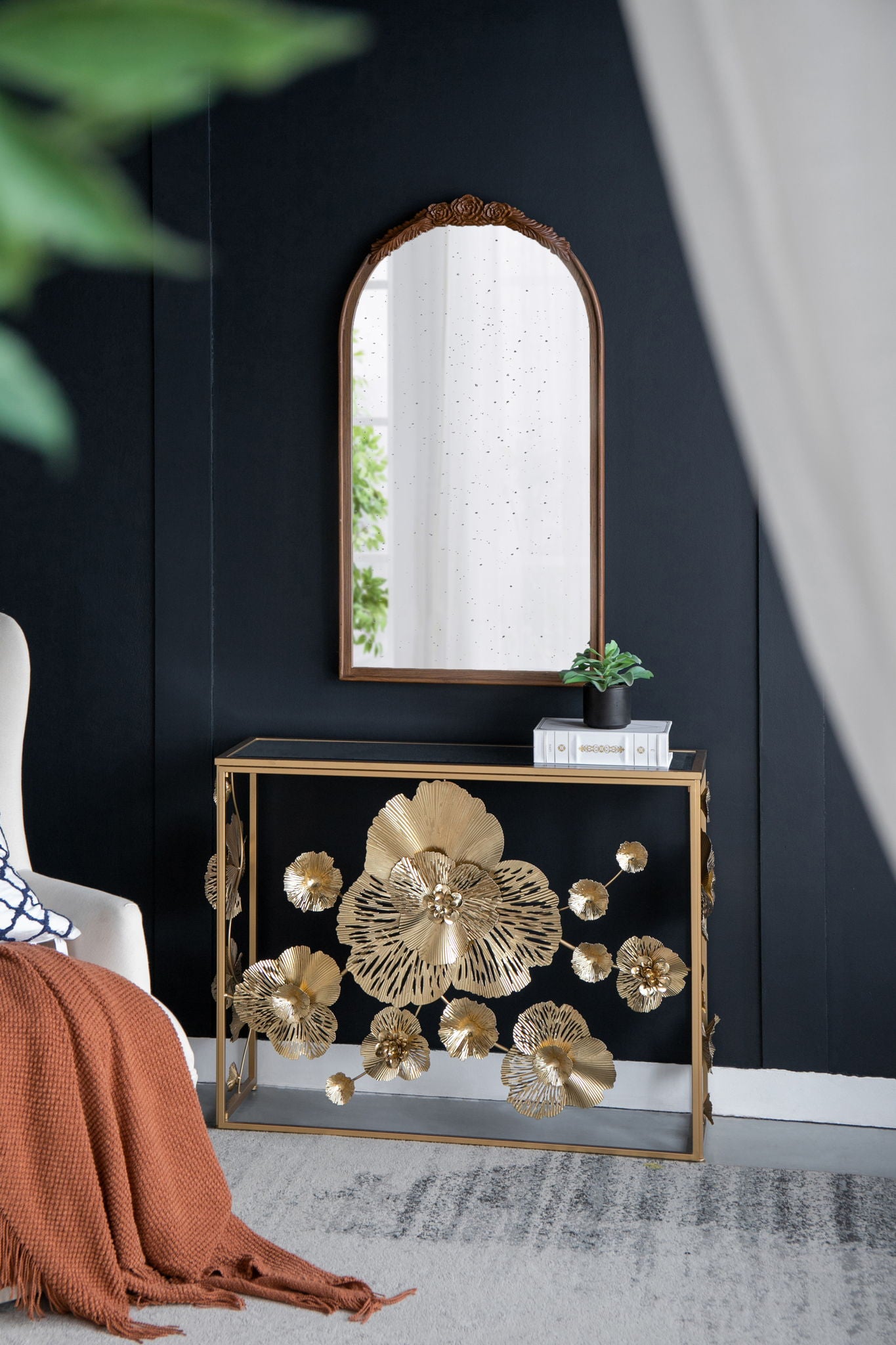 A Console Table With Mirrored Glass Top, Modern Entryway Table For Living Room Foyer Entryway - Gold stands against a dark wall, topped with a plant and books. An arched mirror hangs above, with a white curtain and orange throw blanket nearby.