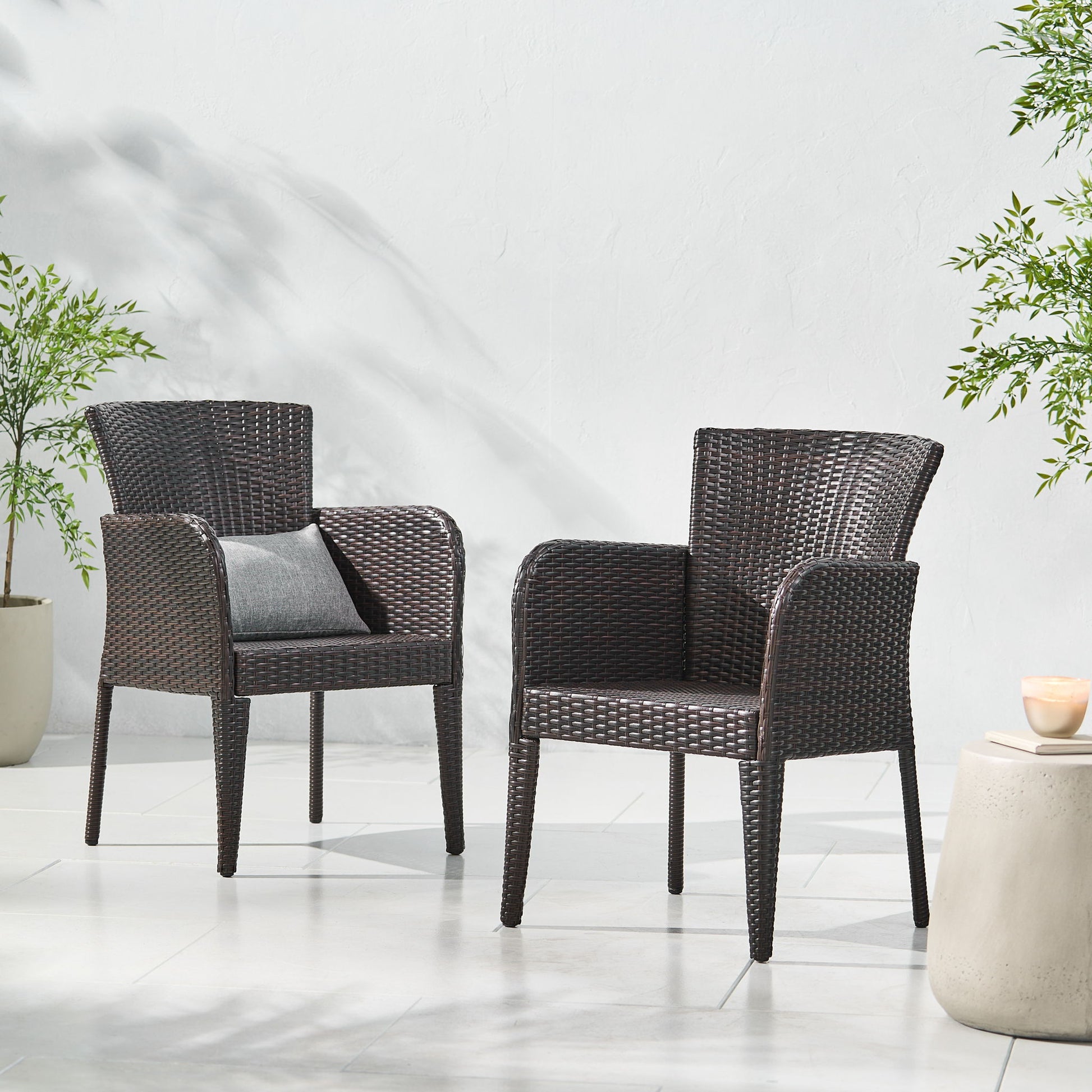 Two Anaya Outdoor Wicker Dining Armchairs sit on a white tiled patio with a pillow on the left chair. Nearby are green potted plants and a small round side table with a candle, set against a light-colored wall.