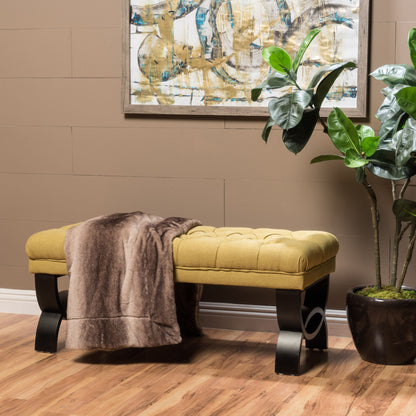 The Hilton - Ottoman Bench Classic Style features a mustard-yellow tufted cushion and black legs, topped with a brown faux fur throw. A potted plant and abstract wall art add warmth to the cozy space on a wooden floor.