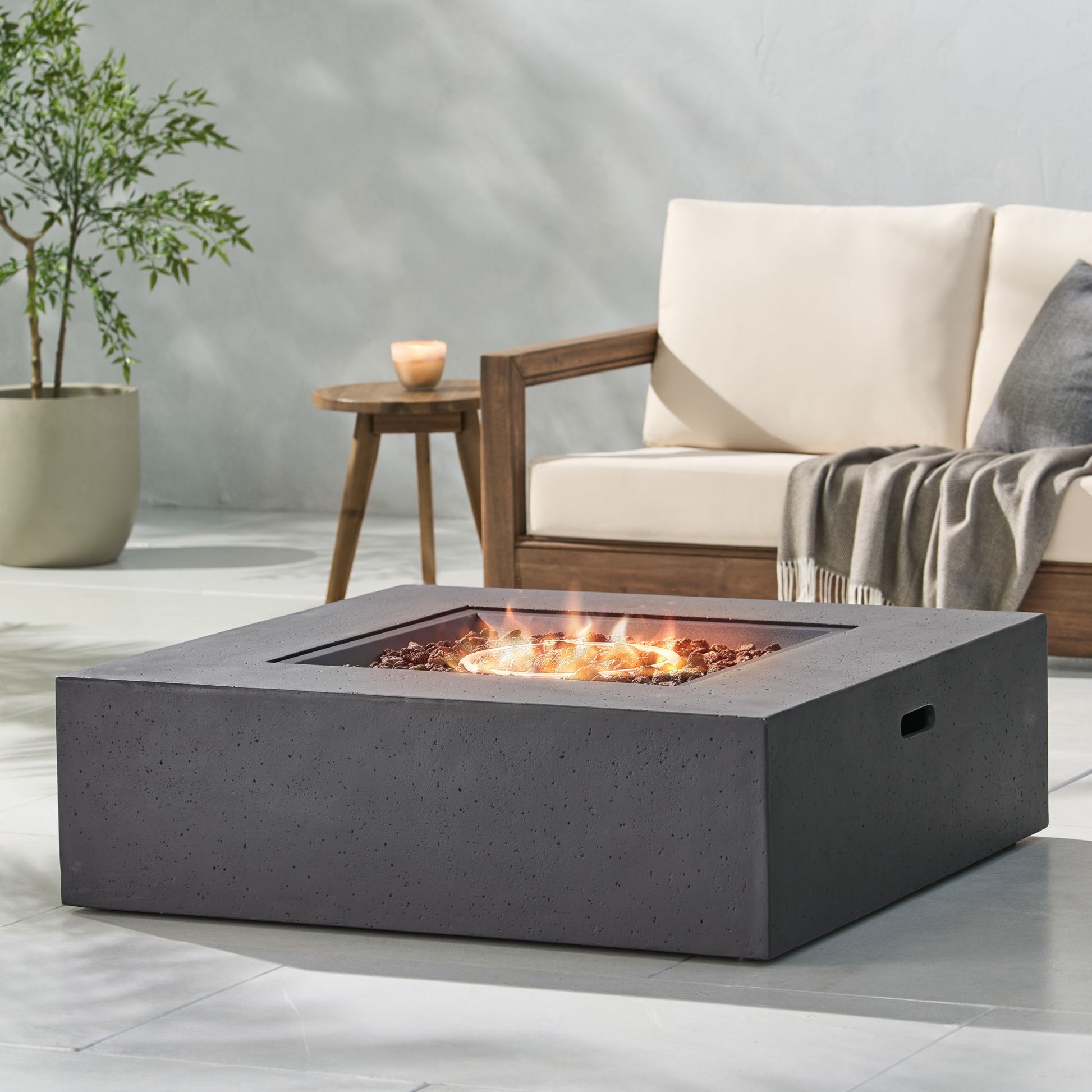 A modern patio featuring the Modern 50,000 Btu Square Fire Table With Propane Tank burning in front, a cushioned wooden sofa with a gray throw blanket behind, and a potted plant on the side.