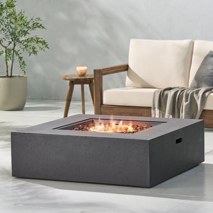 A modern patio featuring the Modern 50,000 Btu Square Fire Table With Propane Tank burning in front, a cushioned wooden sofa with a gray throw blanket behind, and a potted plant on the side.
