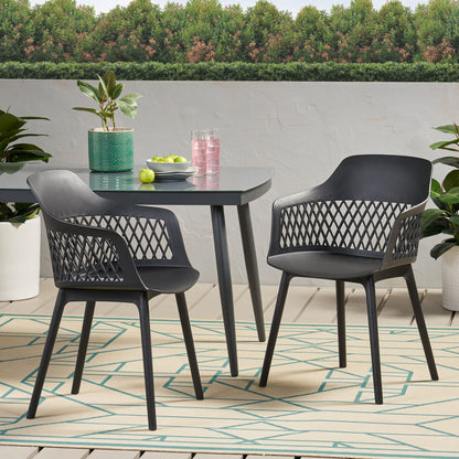 Two Azalea chairs and a table with a plant, green apples, and pink drinks sit on a patio with a geometric rug and potted plants; lush trees are visible in the background.