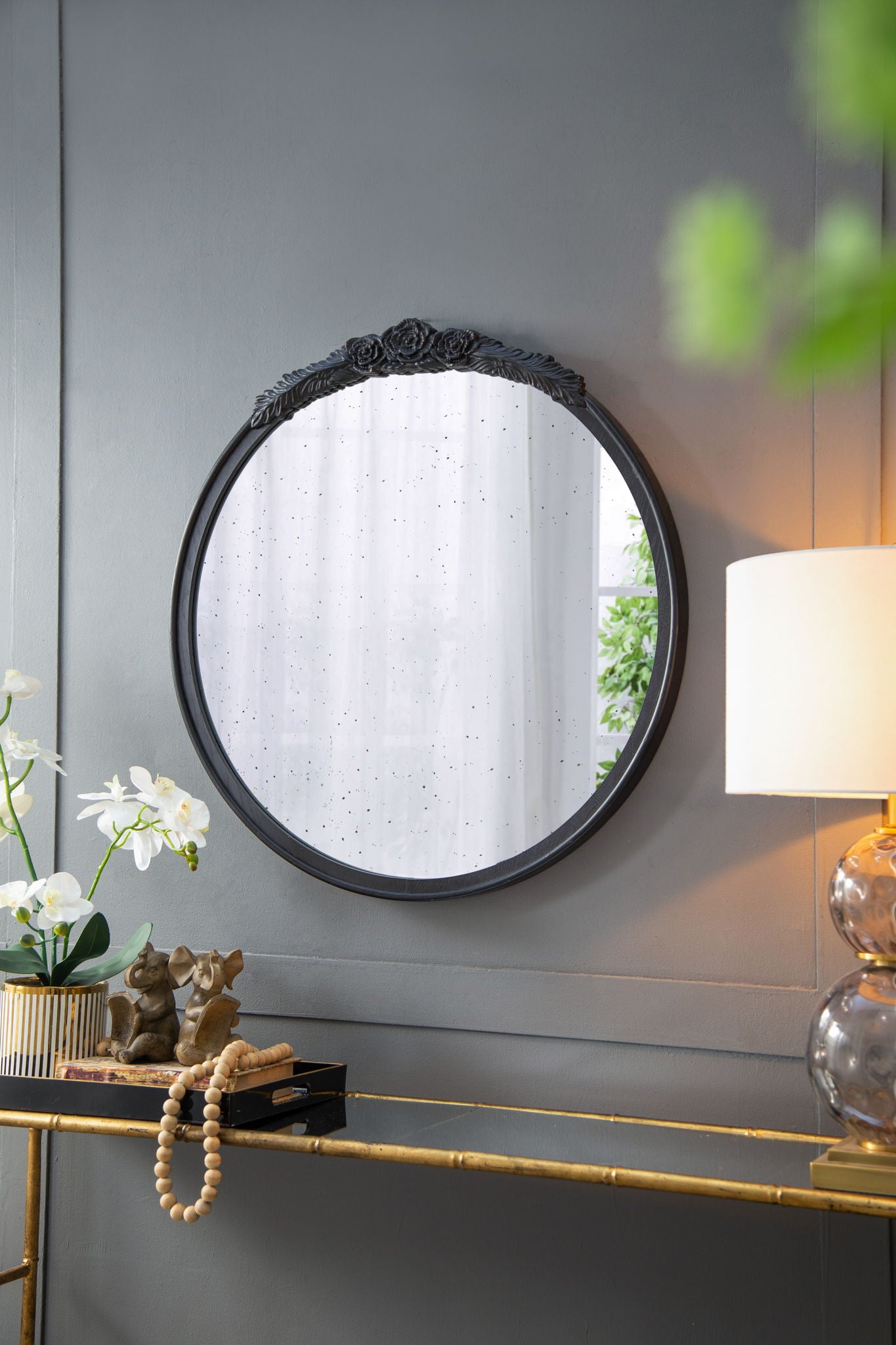 The Round Hand Carved Rose Antique Mirror Frame hangs on a gray wall above a console table with a lamp, orchids, figurines, and books. Sheer curtains and greenery are reflected in the mirror.