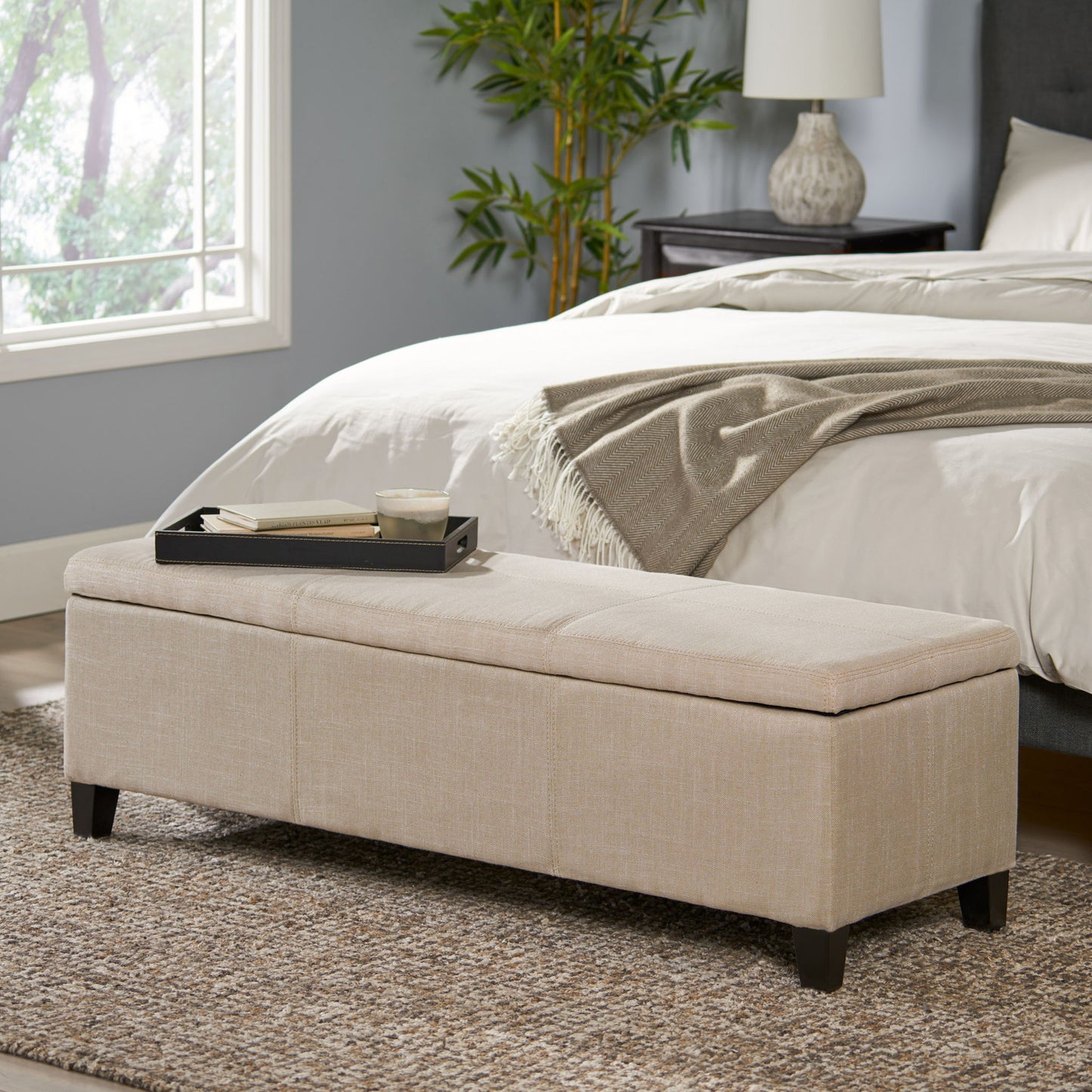 The Glouster Contemporary Storage Ottoman, upholstered in beige, sits at the foot of a bed in a cozy bedroom with gray walls, a large window, a plant, and a nightstand lamp. A tray with a cup and book rests on top.