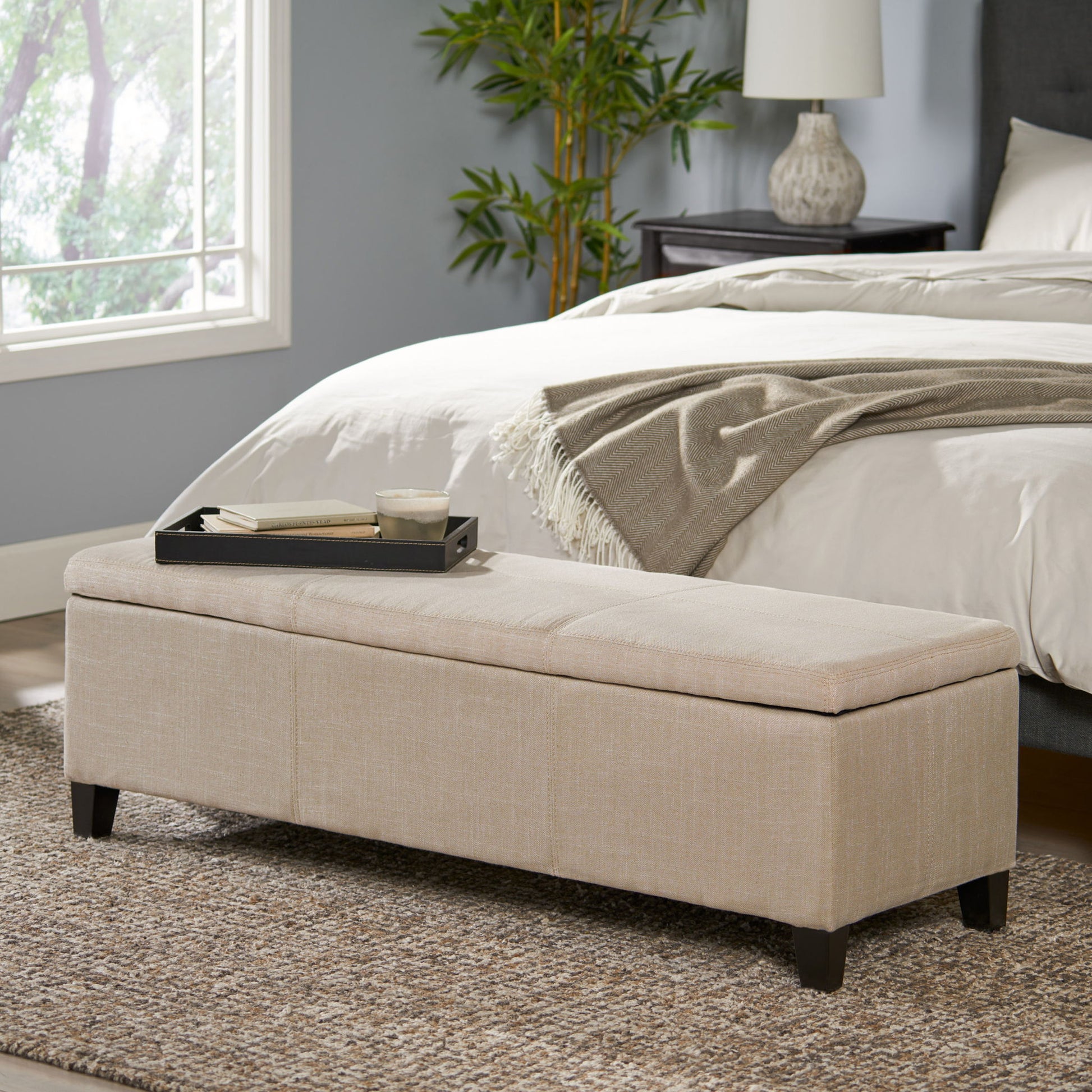 The Glouster Contemporary Storage Ottoman, upholstered in beige, sits at the foot of a bed in a cozy bedroom with gray walls, a large window, a plant, and a nightstand lamp. A tray with a cup and book rests on top.
