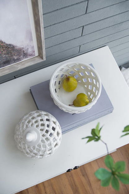 A Ballas - Bird Lidded Jar - White with two pears sits on a closed lavender book atop a white cabinet. The jar’s matching lid is nearby. A framed picture and a green plant are partially visible in the background.