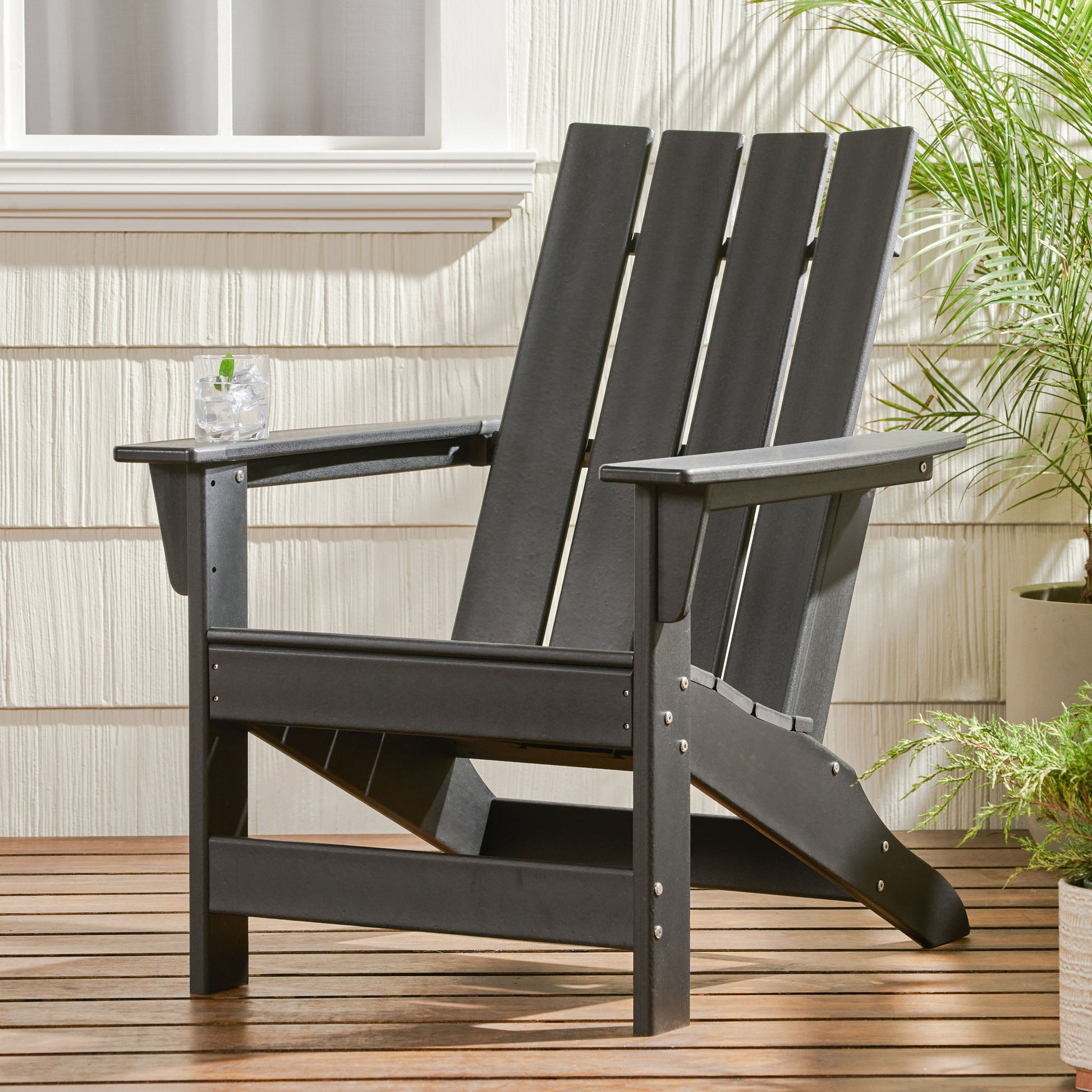 An Enigmara Outdoor High Density Resin Adirondack Chair in black sits on a wooden deck by a white wall with a window, holding a glass of drink on its armrest. Potted green plants are placed nearby for a fresh touch.