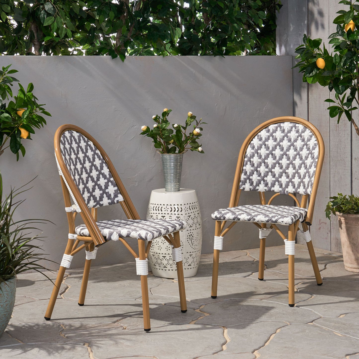 Two Chic French Bistro Chairs with durable rattan and aluminum frames, featuring gray and white cushions, sit on a stone patio with green plants and potted lemon trees nearby. A small white side table with a vase of flowers stands between them.