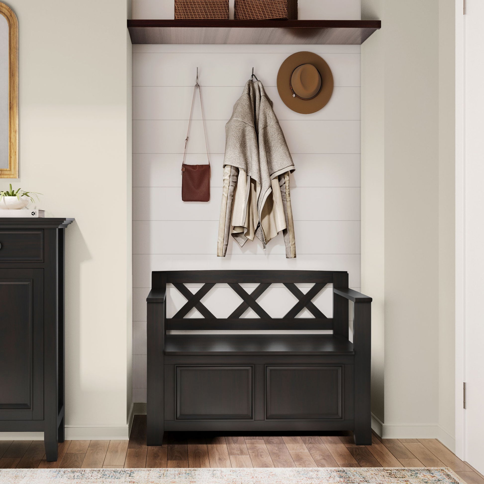 The Amherst - Small Transitional Entryway Storage Bench in black sits against a white shiplap wall. Above, hooks hold a beige coat, brown bag, and tan hat. A shelf with two baskets is mounted above; wooden flooring completes the look.