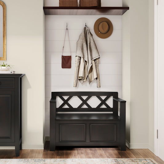 The Amherst - Small Transitional Entryway Storage Bench in black sits against a white shiplap wall. Above, hooks hold a beige coat, brown bag, and tan hat. A shelf with two baskets is mounted above; wooden flooring completes the look.