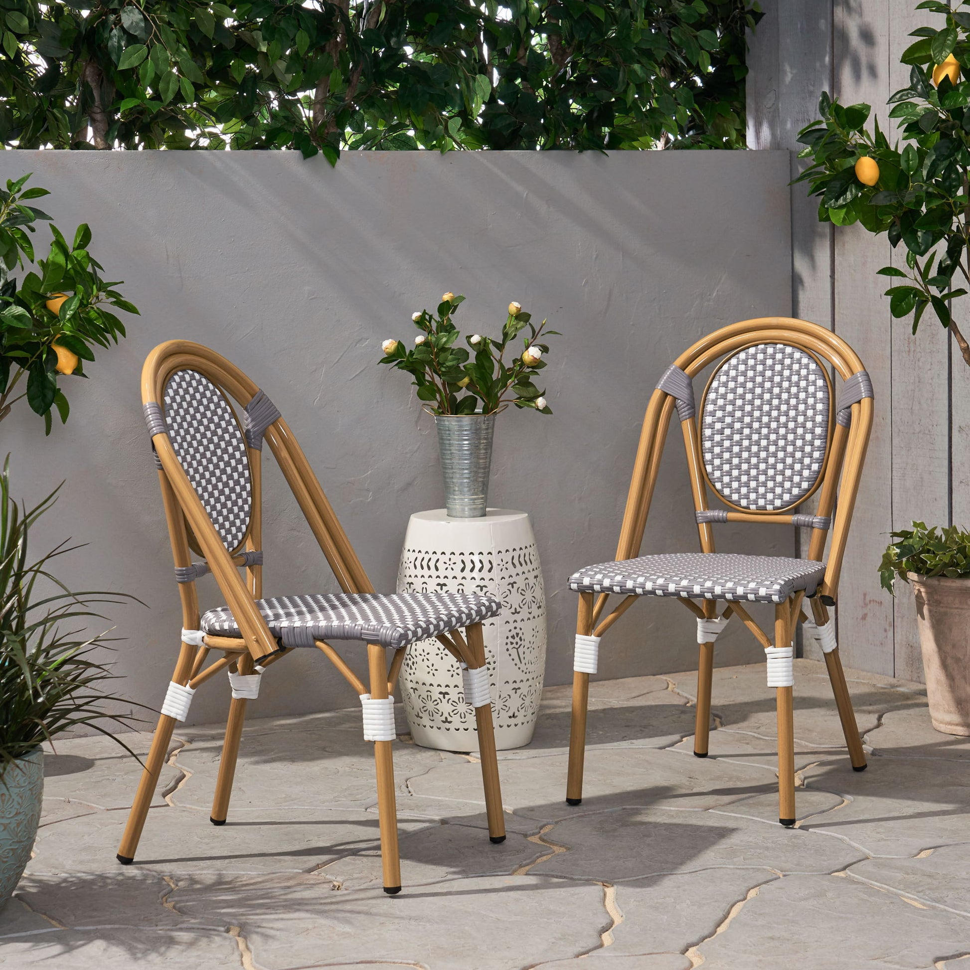 Two Outdoor Rattan And Aluminum French Bistro Chairs with gray and white woven seats sit on a stone patio beside a small white side table with a vase of flowers, surrounded by green plants and a lemon tree.