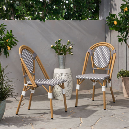 Two Outdoor Rattan And Aluminum French Bistro Chairs with gray and white woven seats sit on a stone patio beside a small white side table with a vase of flowers, surrounded by green plants and a lemon tree.
