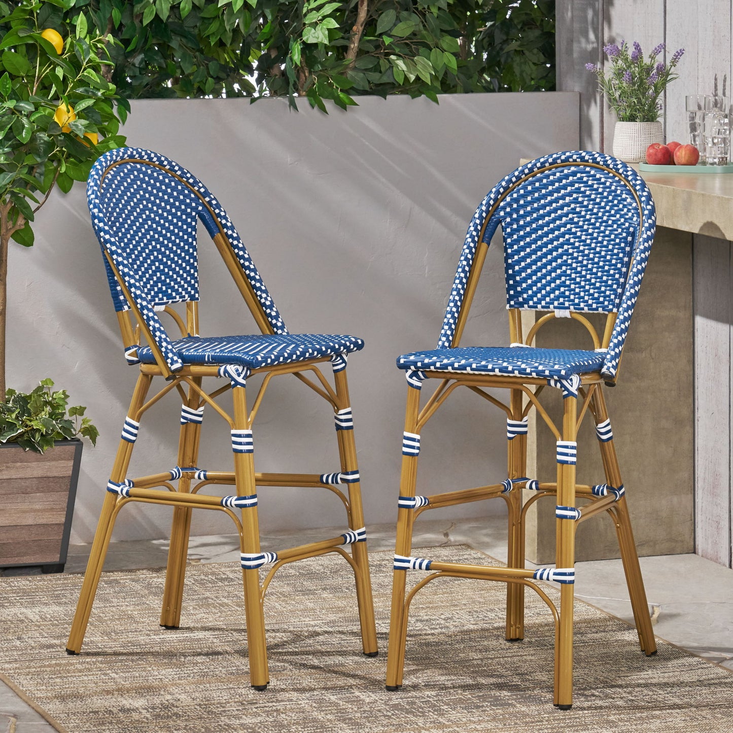 Two Outdoor Pe Rattan French Barstools with blue and white woven seats and bamboo-style legs are placed on an outdoor rug beside a concrete counter topped with potted lavender and apples, surrounded by greenery in the sunlight.