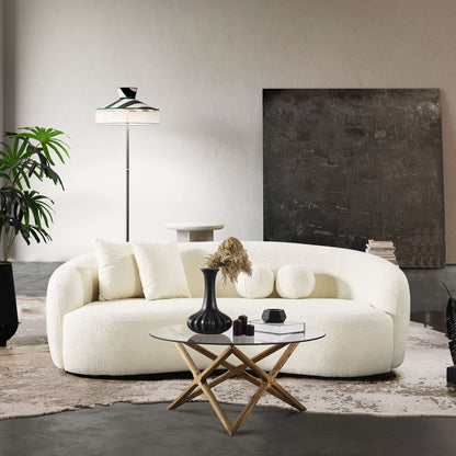 A modern living room featuring the Drake - Japandi Style Curvy Boucle Sofa in white, a round glass coffee table with wooden X-legs, black and white vases, dried flowers, a standing lamp, dark artwork, and green plants on a neutral rug.