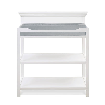 The Universal - Changing Table features a white wooden design with two open shelves below and a gray cushioned changing pad on top, shown against a plain white background.