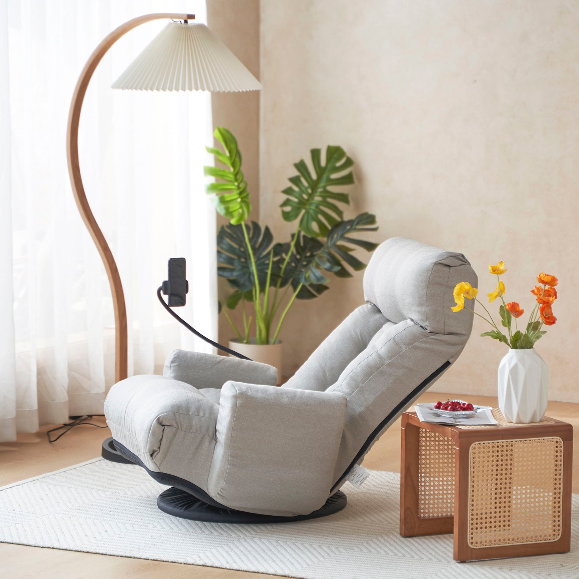 A modern living room featuring an Adjustable Swivel Lounge Chair With 360 Degree Rotating Seat And Headrest Support in light gray, a wooden side table with a vase of yellow-orange flowers, snacks, a curved floor lamp, and green potted plants by a sunlit window.