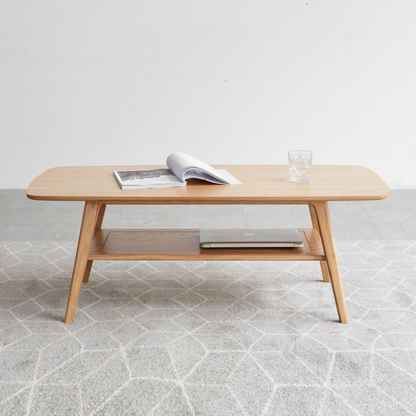 The Low Coffee Table With Storage Shelf displays an open magazine, a glass of water, and a laptop on its lower shelf, set on a light patterned rug against a plain white wall.