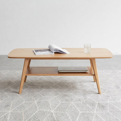 The Low Coffee Table With Storage Shelf displays an open magazine, a glass of water, and a laptop on its lower shelf, set on a light patterned rug against a plain white wall.