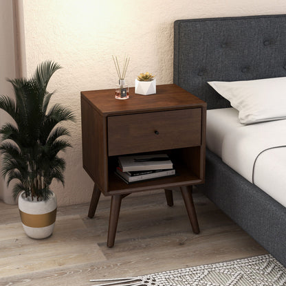 The Caroline Mid-Century Modern Style Night Stand in brown, featuring angled legs, stands beside a gray upholstered bed. On it are a diffuser, candle, and small plant; books are stored below. A potted plant sits nearby on the floor.