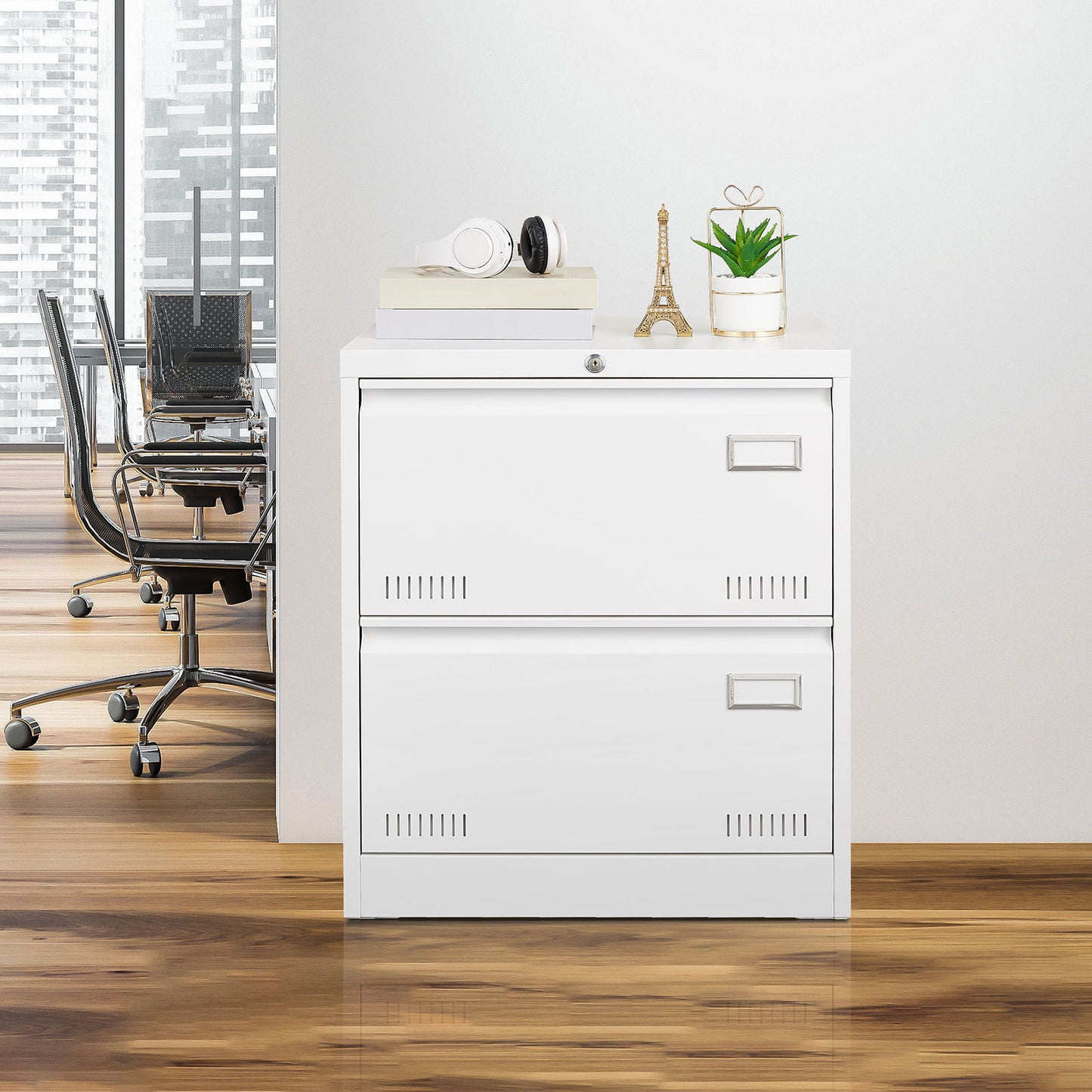 A white Filing Cabinet Lateral File Cabinet 3 Drawer stands against a wall in an office, topped with headphones, a model Eiffel Tower, a plant, and a mug. A glass-walled meeting room with chairs is visible in the background.