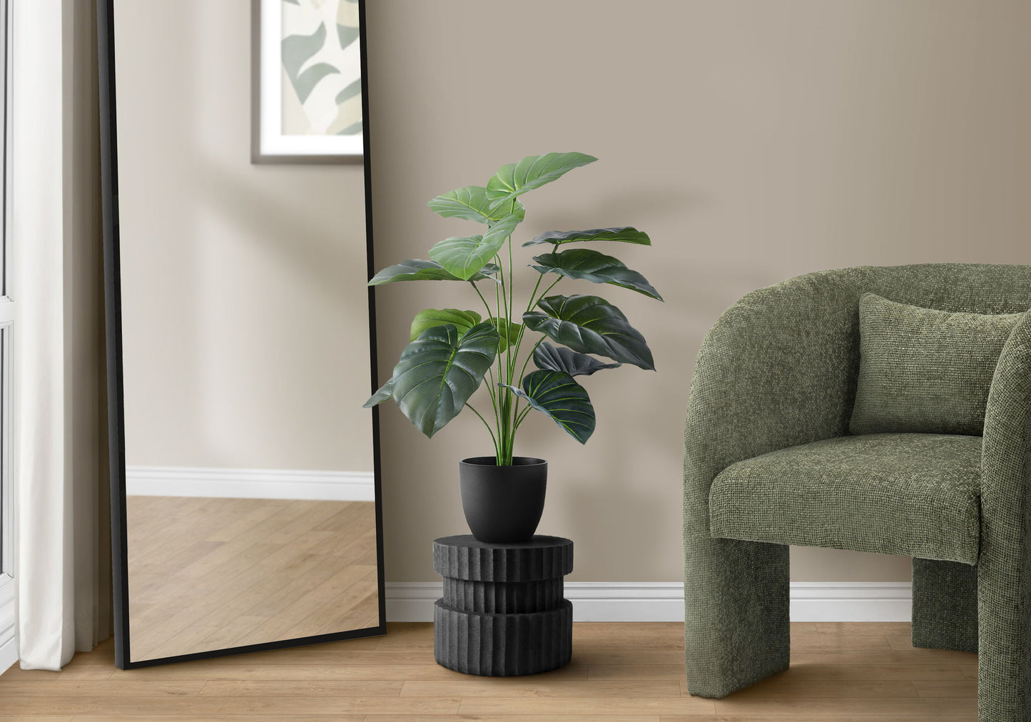 A 24" tall artificial Alocasia plant in a black pot sits on a textured stand next to a large mirror and green armchair in a modern room with wood floors.