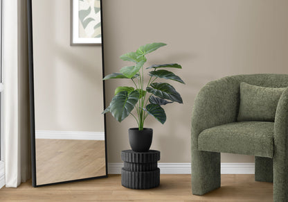 A 24" tall artificial Alocasia plant in a black pot sits on a textured stand next to a large mirror and green armchair in a modern room with wood floors.