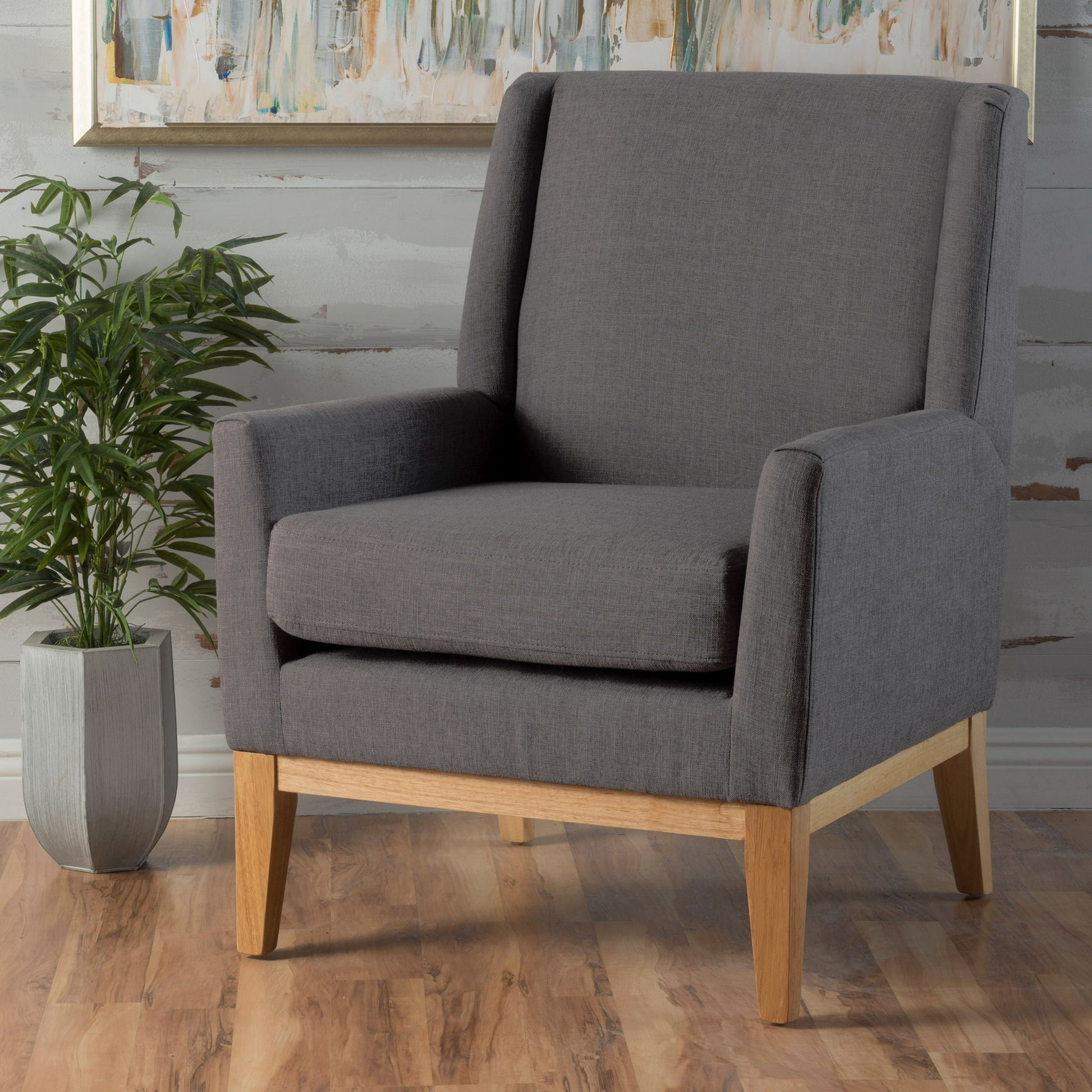 The Modern Accent Chair With Padded Cushion Seat, in gray with wooden legs, sits on a wooden floor near a potted plant and an abstract painting hanging on the wall behind it.