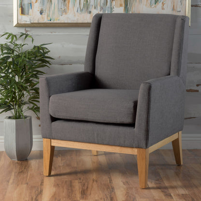 The Modern Accent Chair With Padded Cushion Seat, in gray with wooden legs, sits on a wooden floor near a potted plant and an abstract painting hanging on the wall behind it.