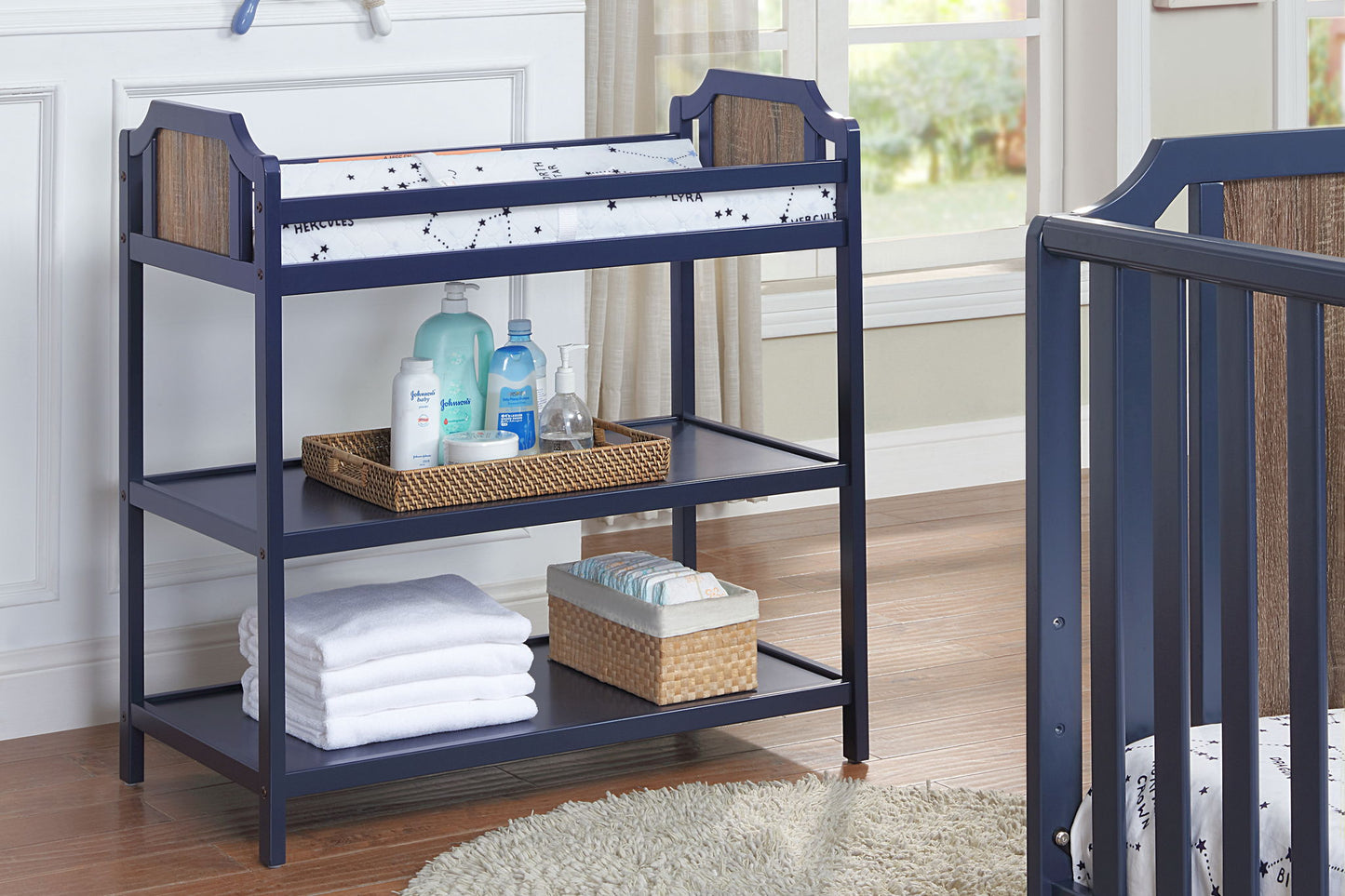 The Brees - Changing Table in navy blue features a cushioned mat, storage baskets with baby essentials, and neatly folded towels on its shelves, set on a wooden floor next to a crib in a softly lit nursery.