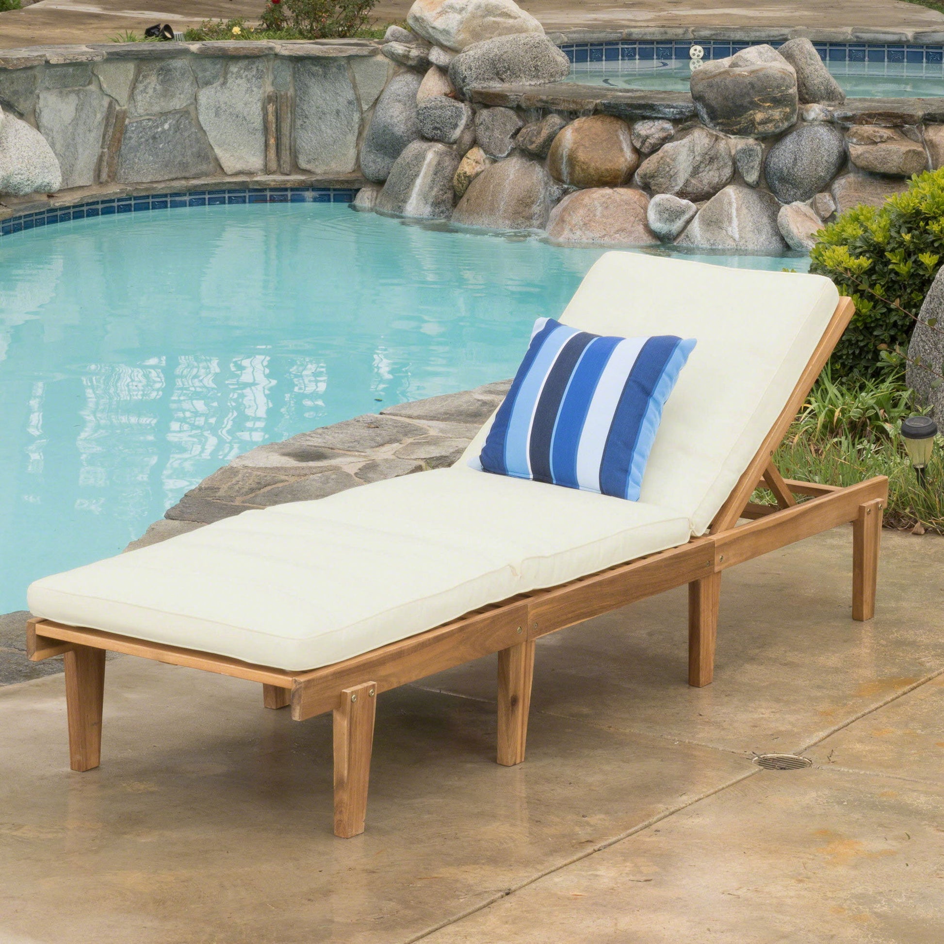 The Ariana Wood Chaise Lounge With Cushion, featuring a blue and white striped pillow, is placed on a concrete patio beside a pool with rock accents.