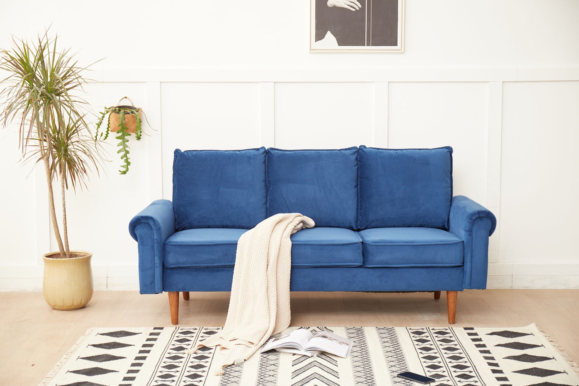 The Graceful Design Plush Comfort Sofa, a blue three-seater, sits against a white wall with a beige blanket over one arm. In front, a potted plant, hanging plant, open book, and smartphone rest on a patterned rug.