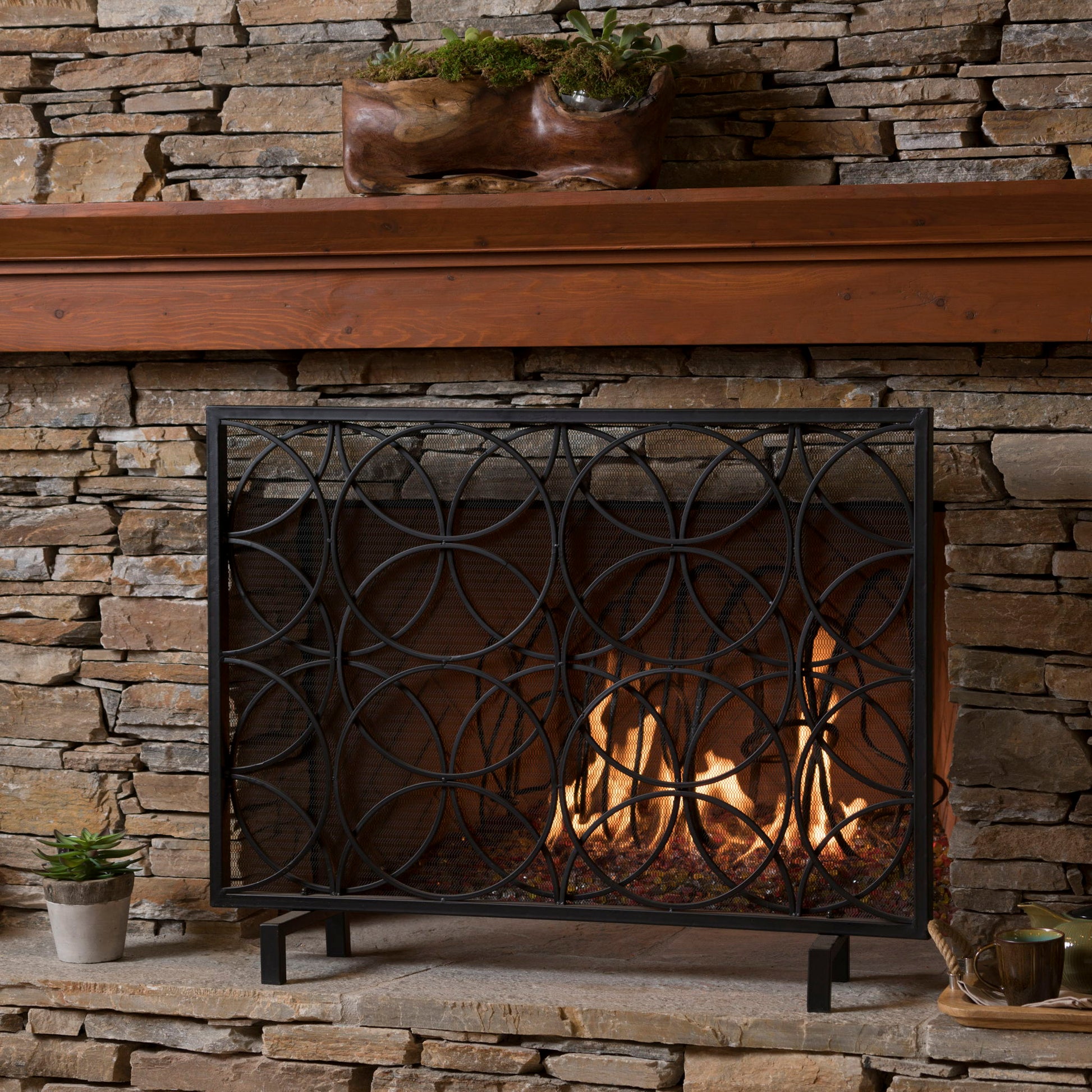 A stone fireplace features a burning fire protected by the Thayer - Single Panel Iron Fireplace Screen With Geometric Design. A plant in a carved wooden planter sits on the mantel, while a small potted plant and cup rest on the hearth.