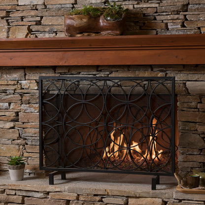 A stone fireplace features a burning fire protected by the Thayer - Single Panel Iron Fireplace Screen With Geometric Design. A plant in a carved wooden planter sits on the mantel, while a small potted plant and cup rest on the hearth.
