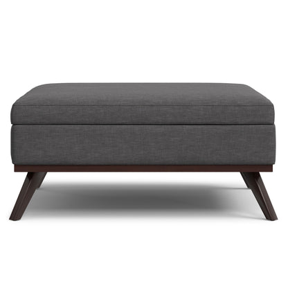 The Owen Multifunctional Square Coffee Table Storage Ottoman features a modern design with dark gray upholstery, a cushioned top, and angled wooden legs, shown from the front against a white background.