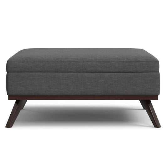 The Owen Multifunctional Square Coffee Table Storage Ottoman features a modern design with dark gray upholstery, a cushioned top, and angled wooden legs, shown from the front against a white background.