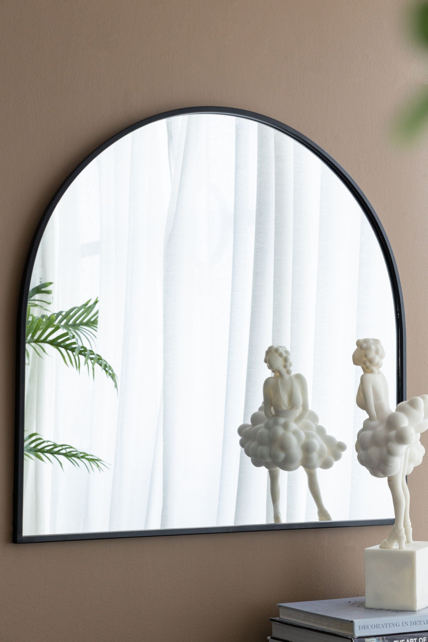 An Arched Mirror With Metal Frame on a beige wall reflects white curtains, a green plant, and a whimsical cloud-like sculpture. Another identical sculpture sits on stacked books nearby. Ideal for living rooms, bedrooms, or entryways.