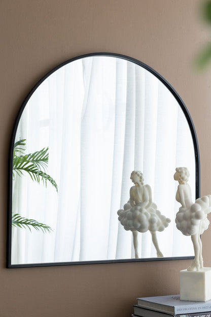 An Arched Mirror With Metal Frame on a beige wall reflects white curtains, a green plant, and a whimsical cloud-like sculpture. Another identical sculpture sits on stacked books nearby. Ideal for living rooms, bedrooms, or entryways.