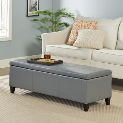 A modern living room features a light sofa, patterned cushions, and the Glouster Contemporary Storage Ottoman in gray used as a coffee table with a tray and drinks, plus a potted plant and large window for natural light.