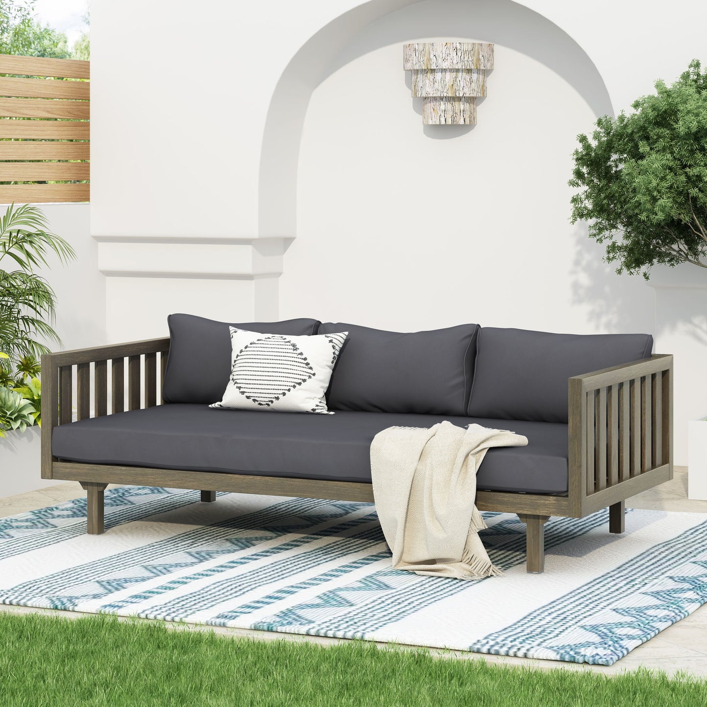 The Claremont 3 Seater Daybed in teak acacia wood with dark gray water-resistant cushions and a striped pillow sits on a blue patterned rug, draped with a cream throw. Green plants and a white wall are in the background.