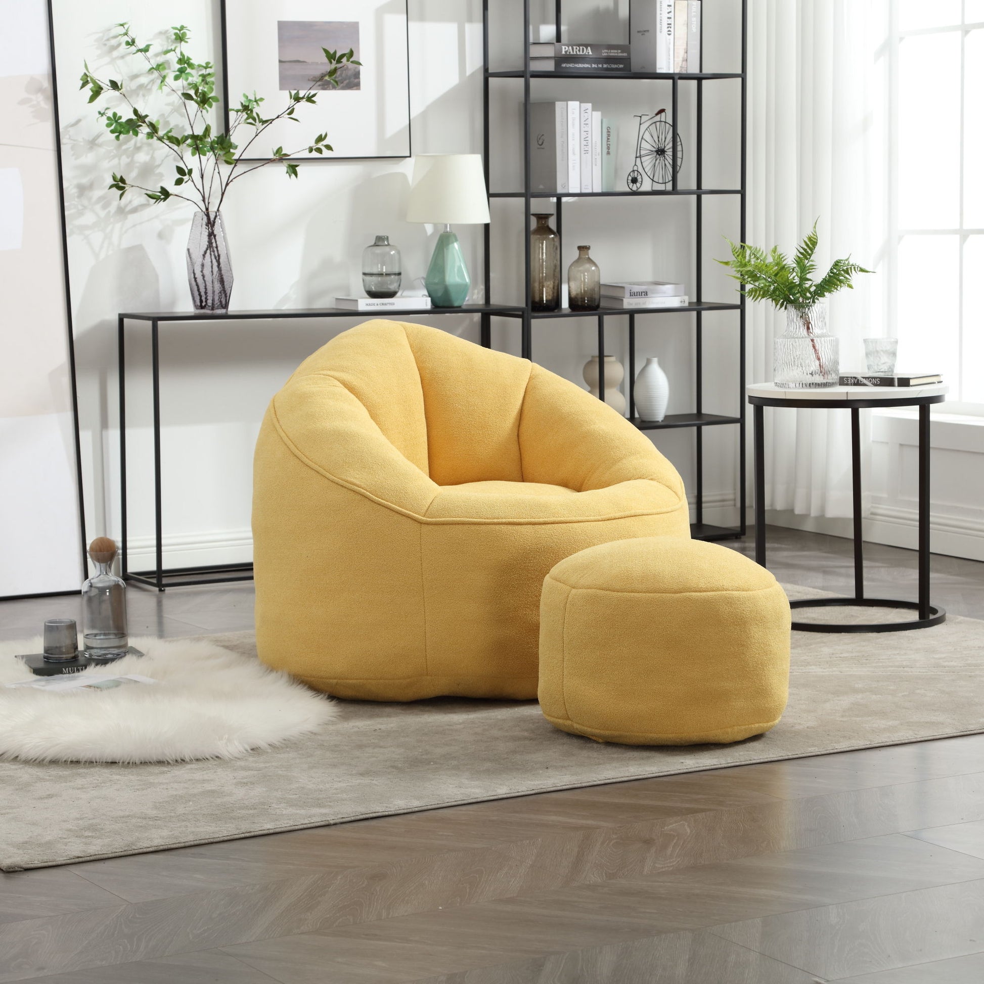 The Bedding Bean Bag Sofa Chair High Pressure Foam with Footrest in yellow sits on a light rug in a modern living room, next to a round side table and a black metal bookshelf decorated with plants and ornaments.