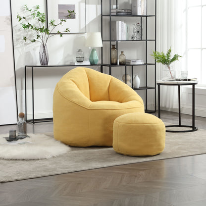 The Bedding Bean Bag Sofa Chair High Pressure Foam with Footrest in yellow sits on a light rug in a modern living room, next to a round side table and a black metal bookshelf decorated with plants and ornaments.
