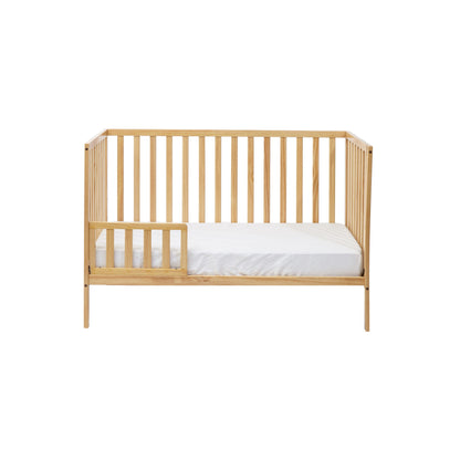 The Palmer - 3-in-1 Convertible Island Crib features a light wood finish, vertical slats on all sides, an open space with a small guard rail on one side, and comes with a white mattress inside.
