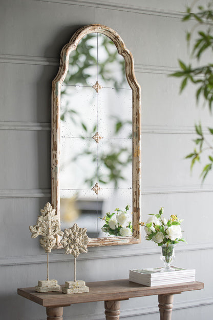 A Large & Framed Wall Mirror, Arched Mirror With Decorative Window Look For Living Room - Cream, hangs above a wooden console table with sculpted ornaments, a clear vase of white flowers, and books against a gray paneled wall.