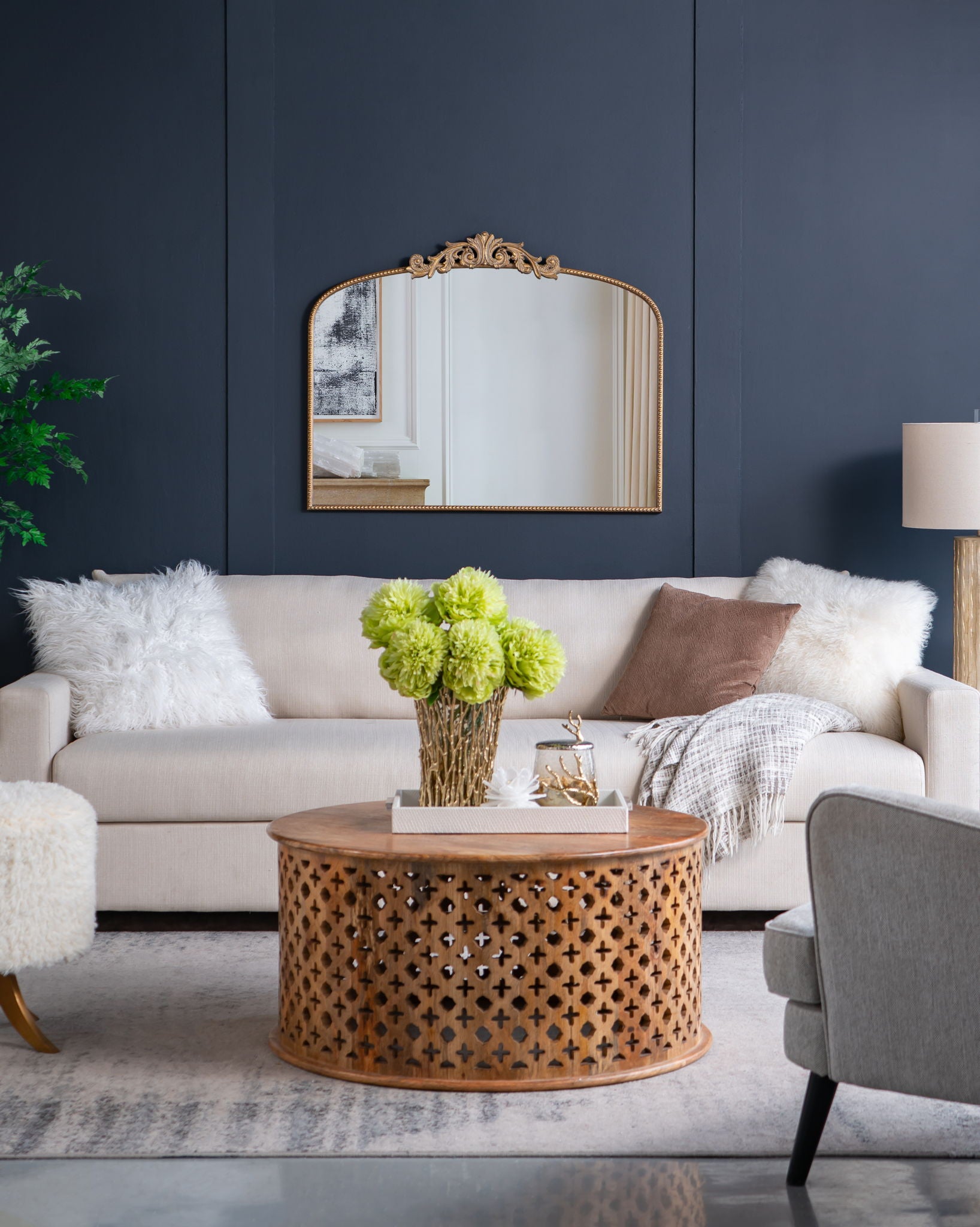 A modern living room with a beige sofa, fluffy white pillows, a brown throw, a round wooden coffee table with cut-out pattern, green flowers in a vase, dark blue walls, and an Arch Mirror—baroque-inspired wall decor for stylish elegance.