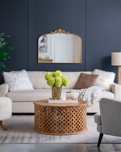 A modern living room with a beige sofa, fluffy white pillows, a brown throw, a round wooden coffee table with cut-out pattern, green flowers in a vase, dark blue walls, and an Arch Mirror—baroque-inspired wall decor for stylish elegance.